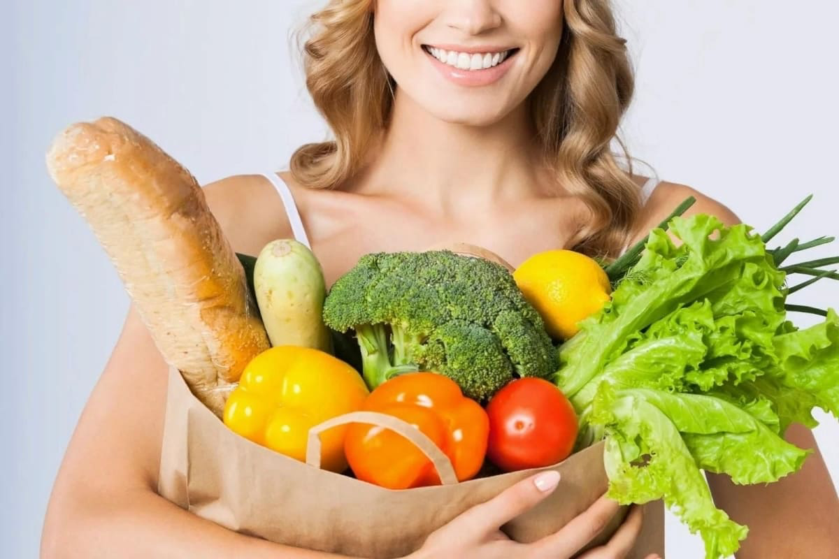 Улыбающаяся женщина держит бумажный пакет с овощами, зеленью и хлебом