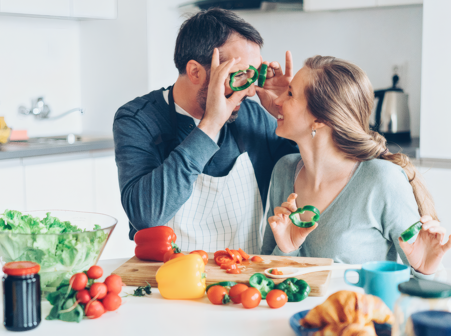 Весёлая пара после 50 лет играет с кольцами зелёного перца на кухне во время приготовления полезного блюда из свежих овощей