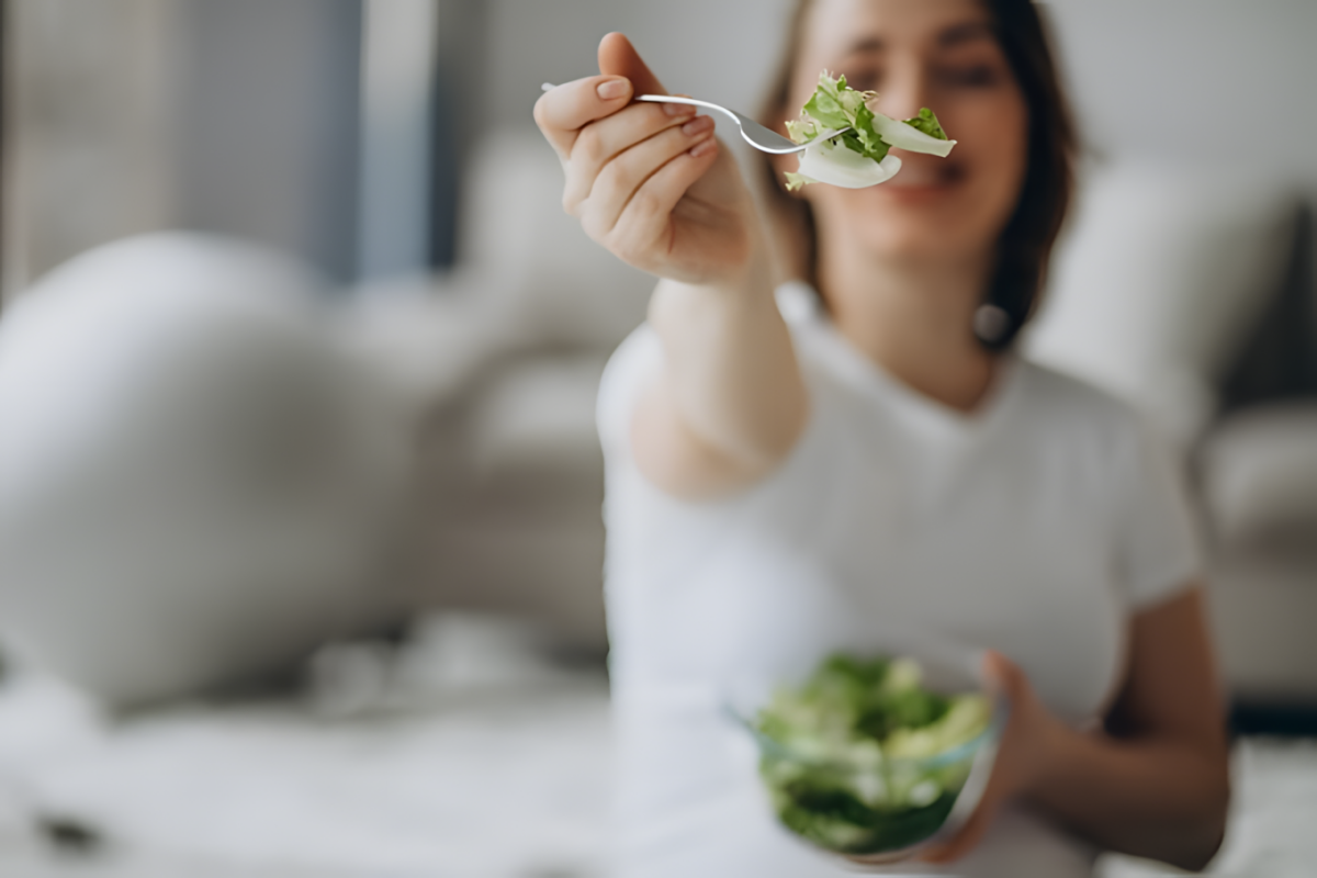 Молодая женщина в белой футболке держит миску с салатом и протягивает вилку с зеленью, приглашая попробовать полезную еду