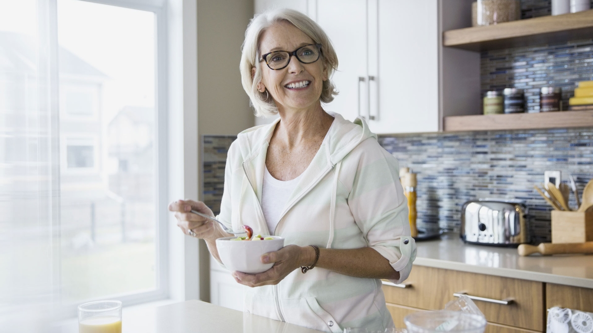 Улыбающаяся женщина после 55 лет в очках ест салат из свежих овощей на кухне